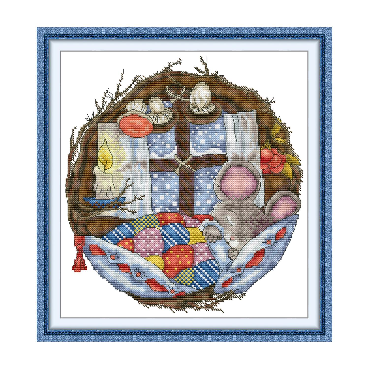 Animal cross stitch design of a sleeping mouse curled in a patchwork quilt inside a round twig frame, with candle and winter berries