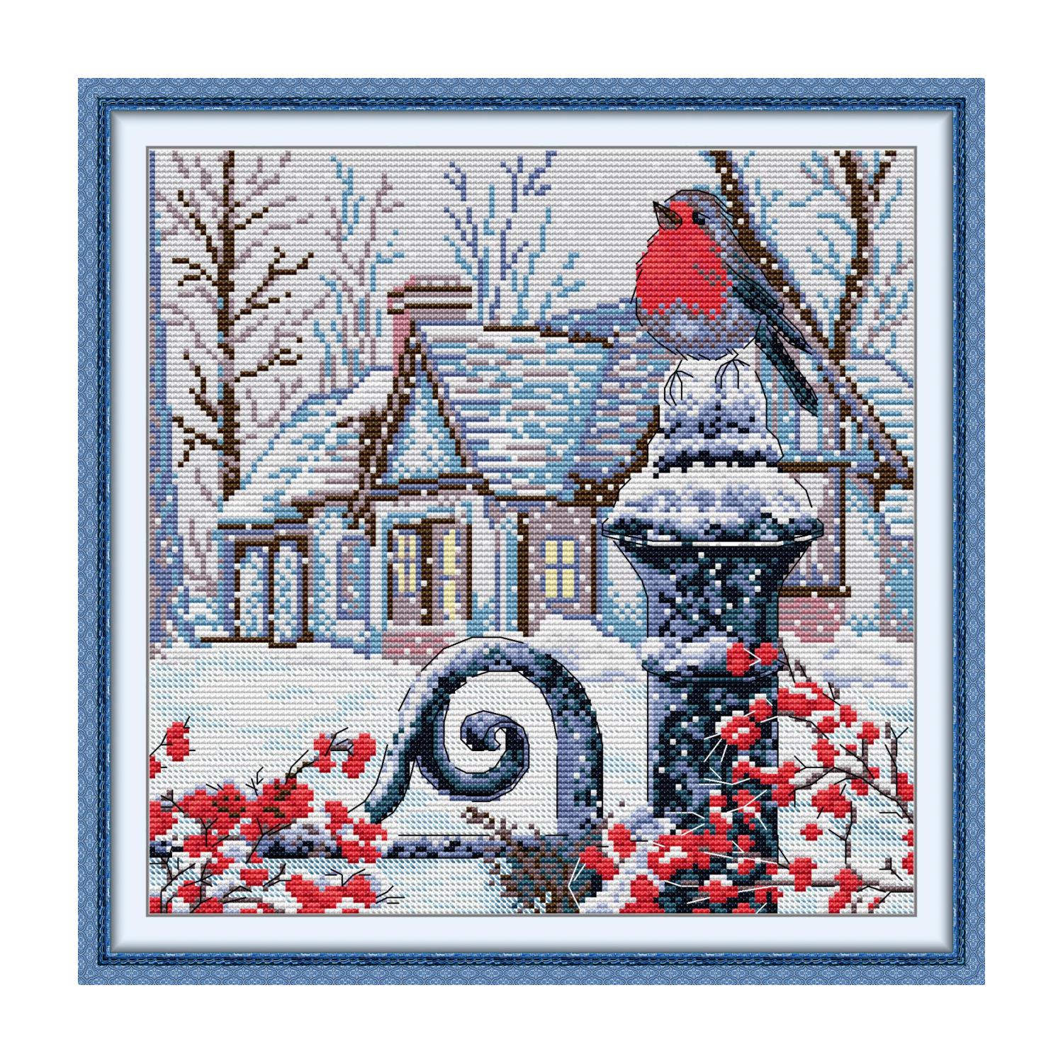 Bird cross stitch design of a robin perched on a snowy birdbath, with a winter cottage and red berry branches in the background